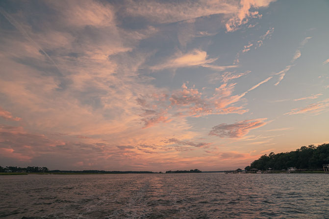 Pink and orange sunset clouds over a coastal estuary, rippling water with a boat wake in the foreground and tree-lined shoreline with docks on the right