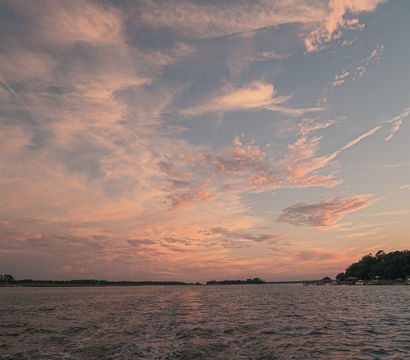 Pink and orange sunset clouds over a coastal estuary, rippling water with a boat wake in the foreground and tree-lined shoreline with docks on the right