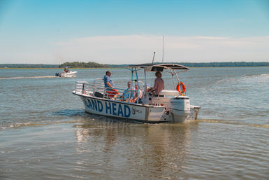 Small tour boat with several people and an outboard motor cruising calm coastal waters near a tree‑lined island under a bright blue sky