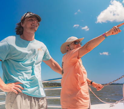 Two men on a sun-soaked boat ride over blue water and sky — one wearing a sun hat and sunglasses points toward the horizon while the other in a light-blue shirt smiles.