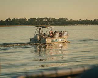 Sunlit sightseeing boat cruising calm waters near Hilton Head Island, SC at golden hour with passengers and tree-lined shoreline reflected in gentle ripples.