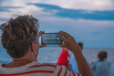 Person on a boat holding a smartphone to capture a cloud-tipped sunset over the calm sea, with blurred passengers and soft blue sky in the background.