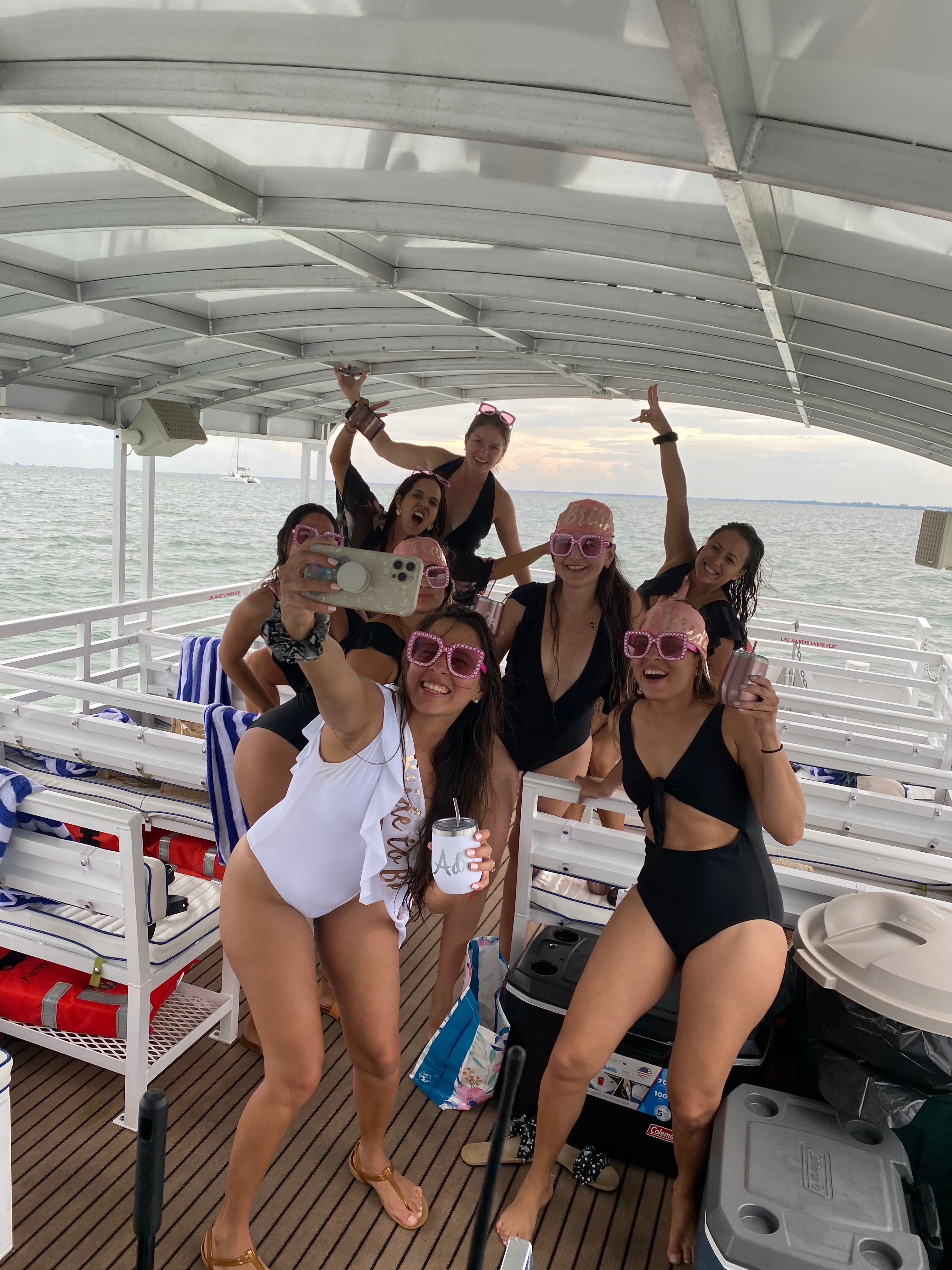 Group of friends in swimsuits taking a selfie on a covered party boat during a coastal sunset cruise, wearing fun pink sunglasses and holding drinks over calm water
