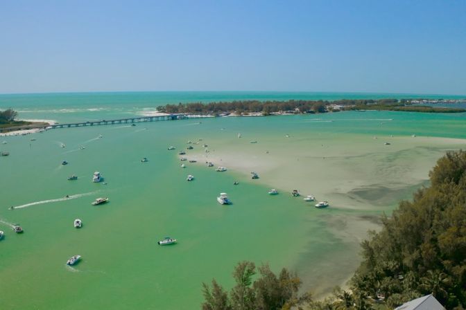 Aerial view of a sunny turquoise bay with dozens of small boats clustered near a sandbar, a low bridge and tree-lined tropical shoreline under a clear blue sky.