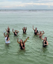 Women in swimsuits and pink sashes raising drinks while standing in shallow green ocean water near a sandy sandbar under a cloudy sky — playful beach bachelorette celebration.