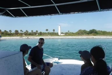 Group on a shaded boat deck looking across turquoise coastal water to a palm-lined sandy beach with a white lighthouse on the shore.
