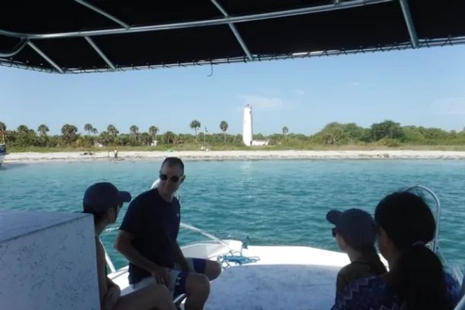 Group on a shaded boat deck looking across turquoise coastal water to a palm-lined sandy beach with a white lighthouse on the shore.