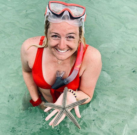 Smiling snorkeler in a red swimsuit with mask and snorkel on her forehead holding a five-armed starfish above clear green tropical water.