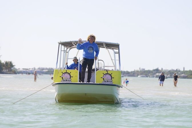 Crew on a small tour boat waving from the bow in clear shallow turquoise water near a sunlit sandbar with people wading and a distant shoreline.