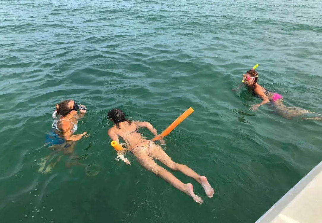 Three snorkelers wearing masks and colorful snorkels float and swim in green open water near a boat, one using an orange pool noodle.