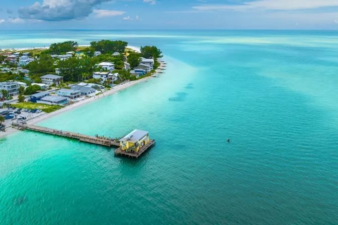 Aerial view of a tropical turquoise coastline with crystal-clear water, a wooden pier leading to a yellow seaside building, white-sand beach and beachfront homes framed by lush greenery.