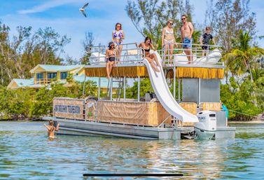Group of family and friends on a two-level tiki-style pontoon boat with a white water slide, sunbathing and swimming in calm tropical coastal waters with palm trees and waterfront homes in the background.
