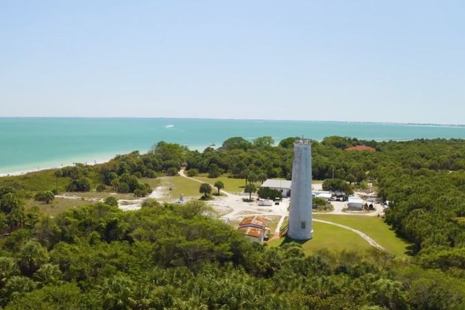 Aerial view of a white lighthouse in a green coastal park overlooking turquoise ocean waters, sandy beach, and clear blue sky