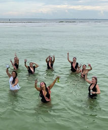Group of women wearing pink sashes and swimsuits toasting and cheering while standing in shallow green ocean water near a calm sandy beach under a cloudy sky.