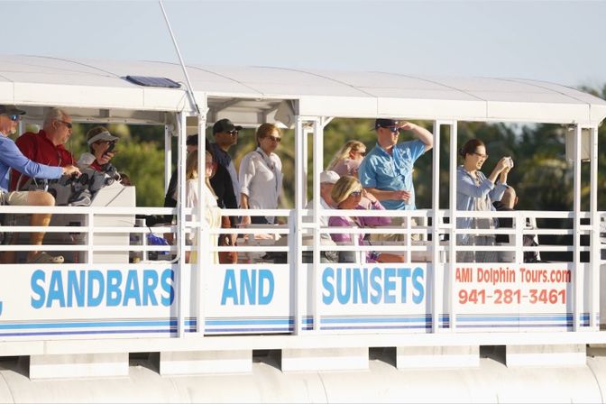 Group of people on a covered coastal sightseeing pontoon boat, some shading their eyes and taking photos while watching the shore, with blue-and-white railing signage visible.