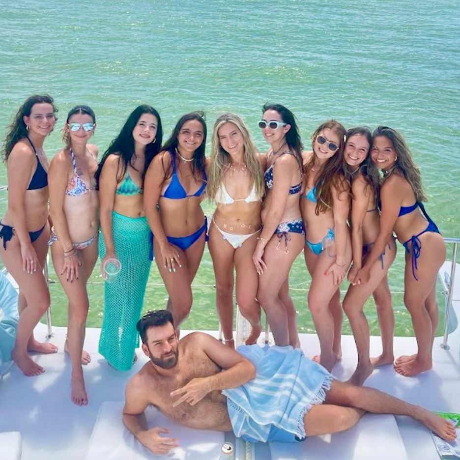 Smiling group of friends in colorful bikinis posing on a boat deck over turquoise water, with a man lounging on a towel in front for a sunny boating party vibe.