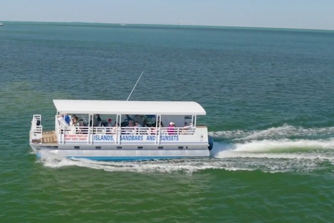 Passenger pontoon tour boat labeled 'Islands, Sandbars and Sunsets' cruising through emerald coastal waters under a white canopy, carrying passengers and leaving a foamy wake.