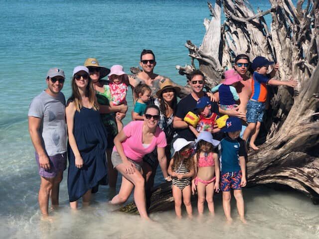 Multi-generation family posing by a large driftwood root on a turquoise tropical beach, adults and children in swimsuits standing in shallow clear water on a sunny day.
