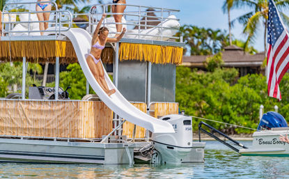 Tiki-decorated pontoon boat with white water slide and woman in a bikini sliding into clear tropical bay, palm trees and American flag in background.