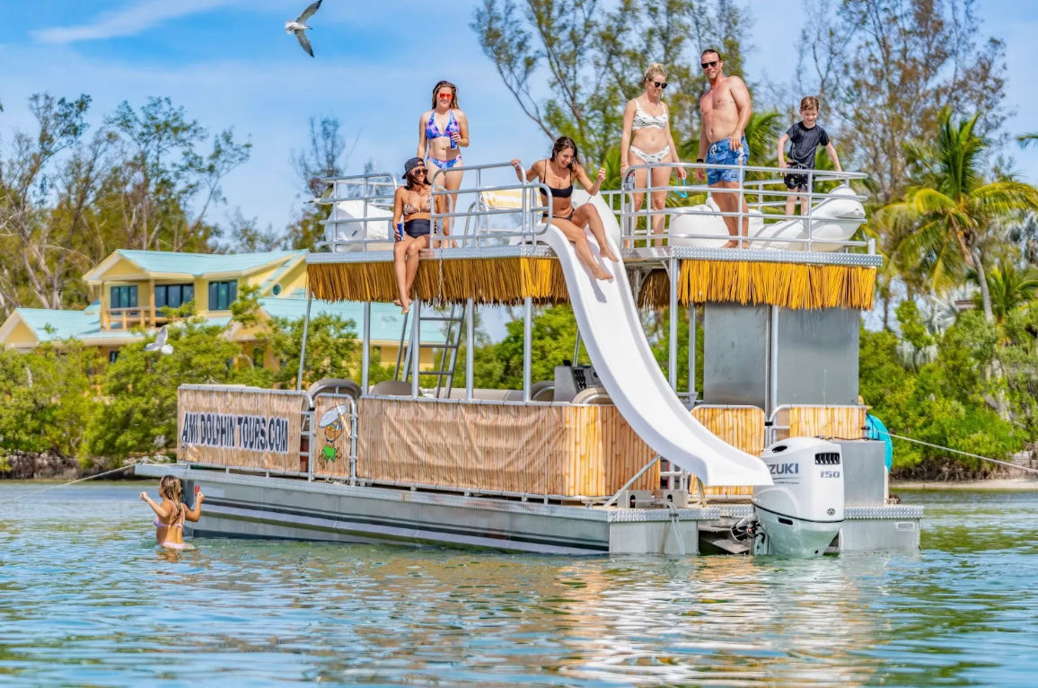 Sun-soaked pontoon boat with a white waterslide and a group of people in swimsuits enjoying shallow tropical water near a palm-lined shoreline, a seagull flying overhead.