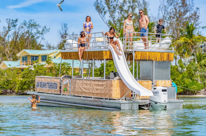 Sun-soaked pontoon boat with a white waterslide and a group of people in swimsuits enjoying shallow tropical water near a palm-lined shoreline, a seagull flying overhead.