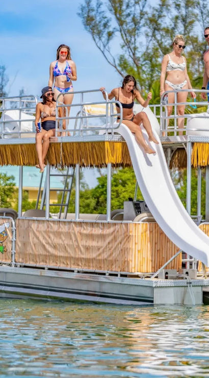 Sun-soaked pontoon boat with a white waterslide and a group of people in swimsuits enjoying shallow tropical water near a palm-lined shoreline, a seagull flying overhead.