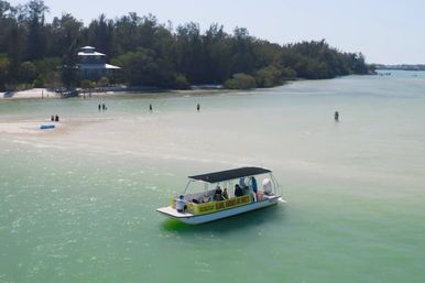 Sunlit covered tour boat in shallow turquoise water near a white sandbar and tree-lined shoreline, with a few people wading and relaxing on the beach