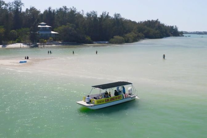 Sunlit covered tour boat in shallow turquoise water near a white sandbar and tree-lined shoreline, with a few people wading and relaxing on the beach