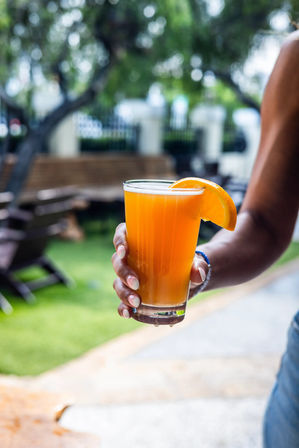 Hand holding a tall glass of bright orange drink garnished with an orange slice on a sunny backyard patio with blurred green lawn and wooden benches