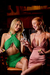 Two friends at a dimly lit cocktail bar laughing and toasting with a salt-rimmed shot glass, one wearing a green shell‑trimmed crop top and skirt and the other in a pink satin dress holding lime wedges