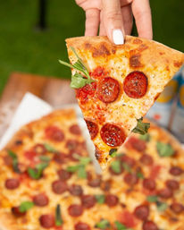 Hand with white nail polish lifting a pepperoni pizza slice topped with melted cheese and fresh arugula from a whole pie on a wooden table outdoors