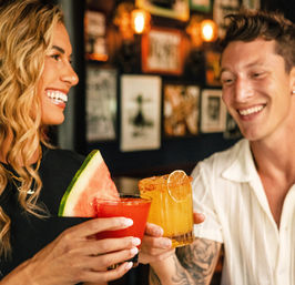 Smiling friends clinking colorful cocktails at a cozy bar — watermelon‑garnished red drink and chili‑rimmed orange cocktail with lime, warm lights in background.