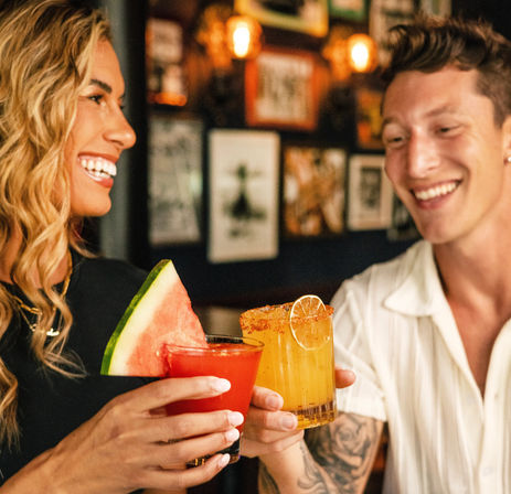 Smiling friends clinking colorful cocktails at a cozy bar — watermelon‑garnished red drink and chili‑rimmed orange cocktail with lime, warm lights in background.