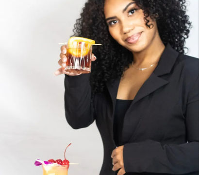 Smiling woman in a black blazer holding up a rocks glass with an orange peel garnish, presenting a lineup of colorful cocktails (tropical hurricane with cherries, blue martini, pink drink) on a white background.