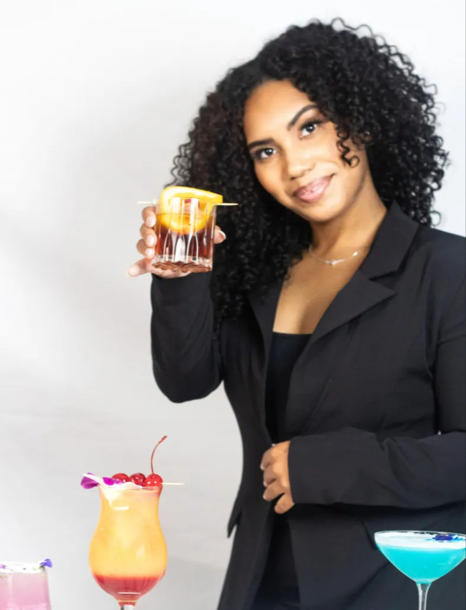 Smiling woman in a black blazer holding up a rocks glass with an orange peel garnish, presenting a lineup of colorful cocktails (tropical hurricane with cherries, blue martini, pink drink) on a white background.
