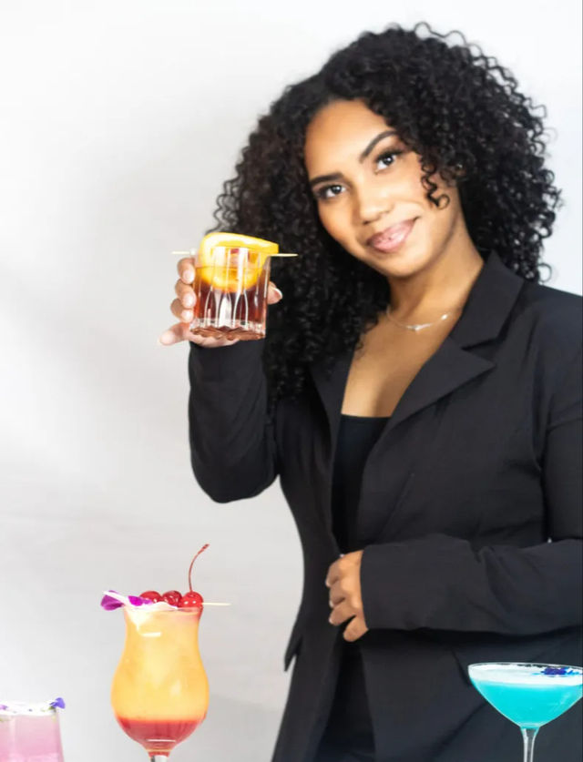 Smiling woman in a black blazer holding up a rocks glass with an orange peel garnish, presenting a lineup of colorful cocktails (tropical hurricane with cherries, blue martini, pink drink) on a white background.