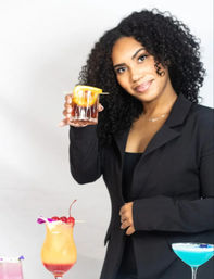 Smiling mixologist in a black blazer holding an Old Fashioned with an orange peel, presenting colorful cocktails on a bar table (blue martini, layered tropical drink with cherry).