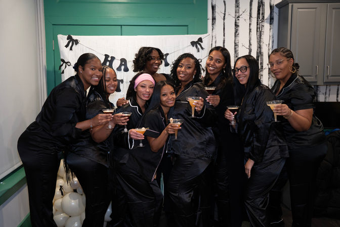 Group photo of nine women in matching black satin pajamas smiling and raising cocktails at an indoor pajama party celebration with balloons and a festive banner.