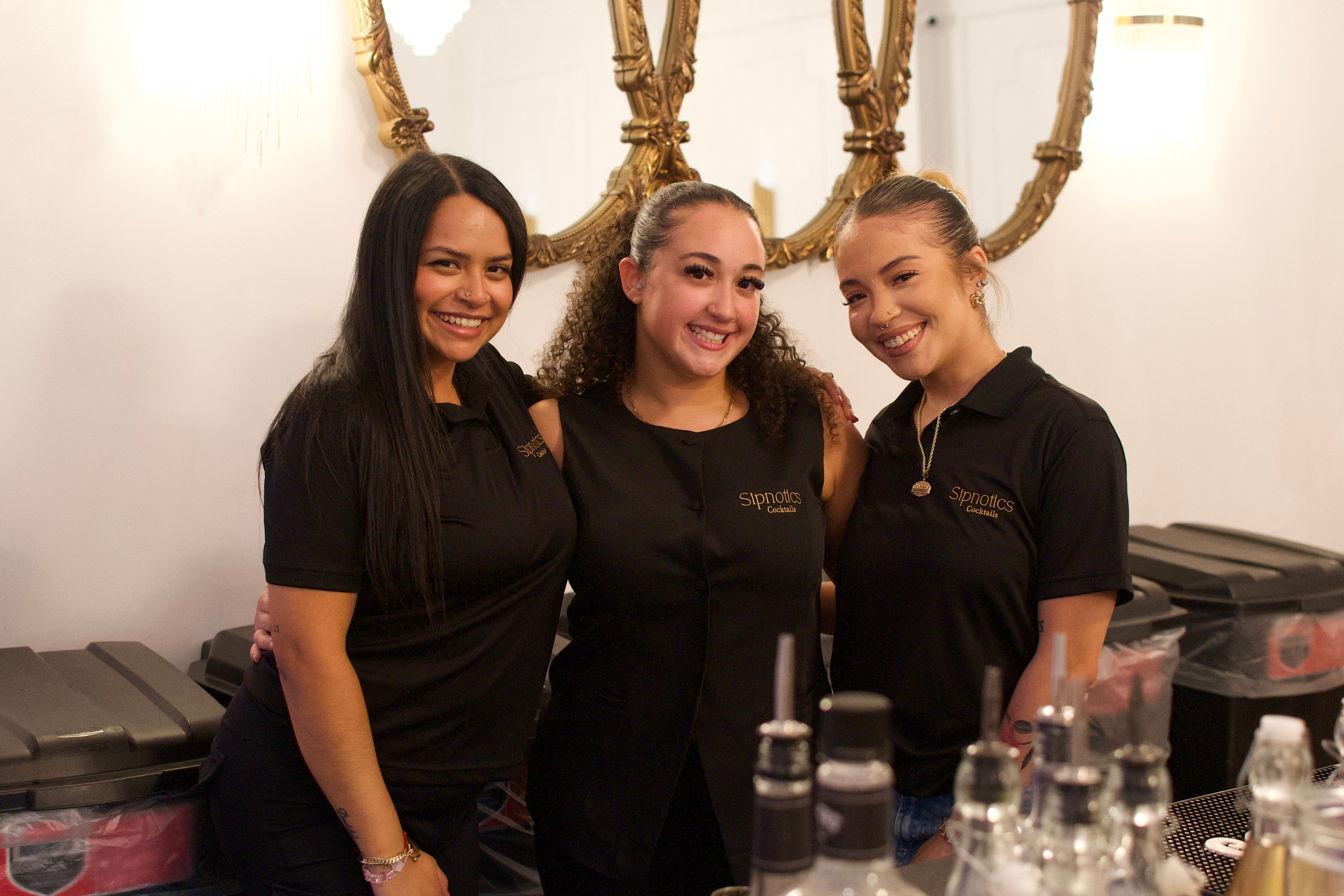 Three smiling bartenders in matching black shirts pose behind a bar with bottles and ornate gold mirrors on a white wall.