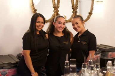 Three smiling bartenders in matching black shirts pose behind a bar with bottles and ornate gold mirrors on a white wall.
