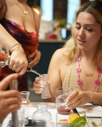 Close-up of two people making cocktails indoors — one pours clear liquor into a jigger held by the other, with a mixing glass, shaker, sliced orange and herbs on the table.