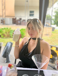Woman in a black halter top holding a cocktail shaker with a creamy drink at an outdoor patio bar, martini glass and fruit skewer on the table, sunglasses perched on her head.
