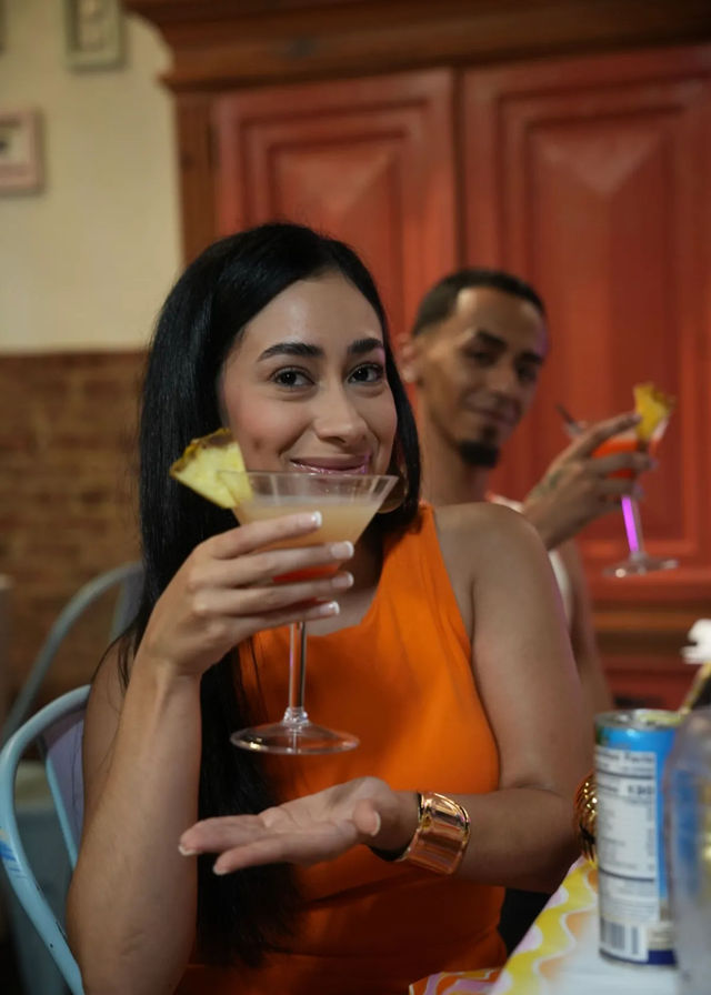 Smiling woman in an orange dress holding a martini-style cocktail with pineapple garnish in a cozy bar, friend toasting in the background.