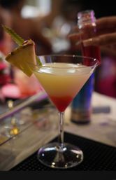 Tropical layered cocktail in a martini glass with pineapple wedge garnish and red syrup sinking to the bottom, blurred bar background