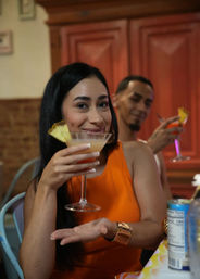 Smiling woman in an orange dress raises a pineapple-garnished martini at a cozy indoor bar or restaurant, with a friend in the background toasting with a cocktail.