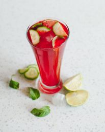 Refreshing bright red iced drink in a tall glass garnished with cucumber slices and mint; lime wedges, cucumber rounds and mint leaves scattered on a speckled white countertop.