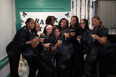 Group photo of nine women in matching black satin pajamas smiling and raising cocktails at an indoor pajama party celebration with balloons and a festive banner.
