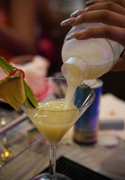 Close-up of a frothy tropical cocktail being poured into a martini glass at a bar, garnished with a pineapple wedge and cherry, with a manicured hand holding the shaker.