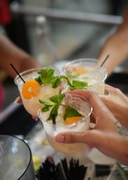Four refreshing summer cocktails in clear plastic cups garnished with fresh mint and peach slices, black straws, hands clinking in a casual toast.