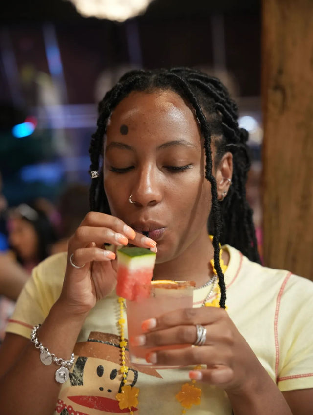 Person with braided hair and nose ring sipping a pink watermelon cocktail garnished with a watermelon cube and orange-rimmed glass in a lively bar setting.